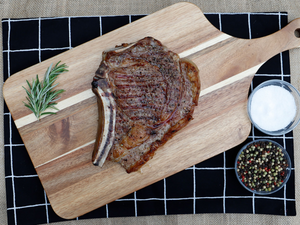Grilled, Bone In Beef Ribeye on a cutting board with green herbs and bowls of salt and pepper.